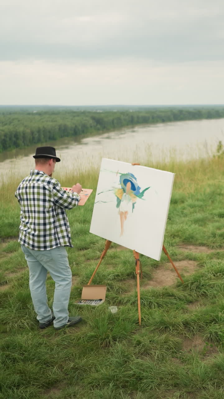 A skilled man, in a hat, checkered shirt, and jeans, is focused on creating a masterpiece on a canvas in a lush grassy field beside a lake. a woman in a hat and white dress sits comfortably on a chair