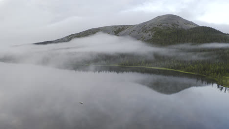 Vista-Aérea-De-Nubes-Bajas-Sobre-Un-Lago-Con-Un-Bote-Pequeño-Y-Una-Montaña-En-El-Fondo