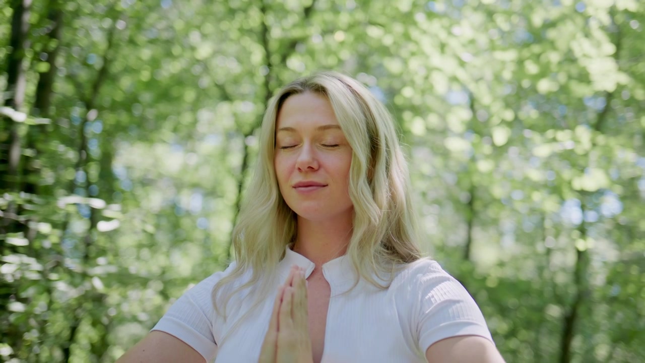 Una mujer medita pacíficamente en un bosque iluminado por el sol. Cierra los ojos y coloca sus manos en posición de oración. Su expresión tranquila muestra serenidad interior. Ideal para contenido de bienestar, atención plena o naturaleza.
