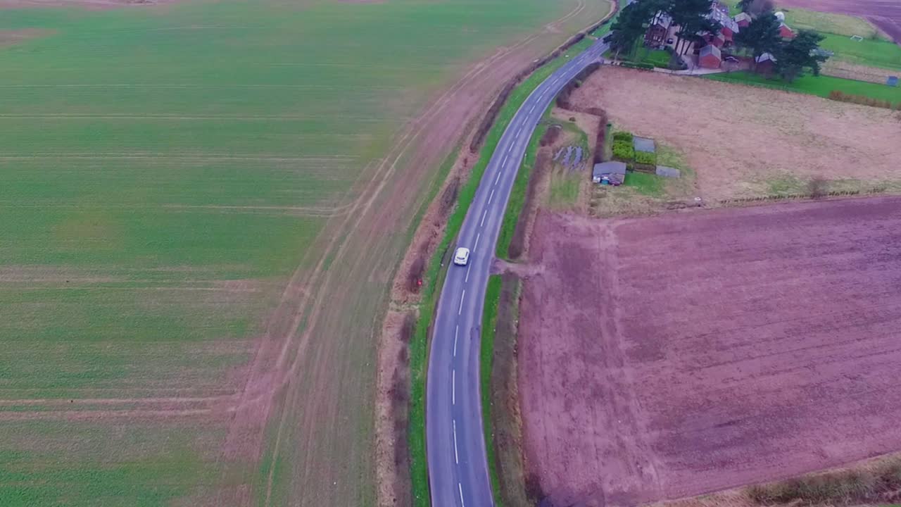 Drone tracking shot of white car driving along road through farmland. Filmed in winter, showing boggy agricultural pasture