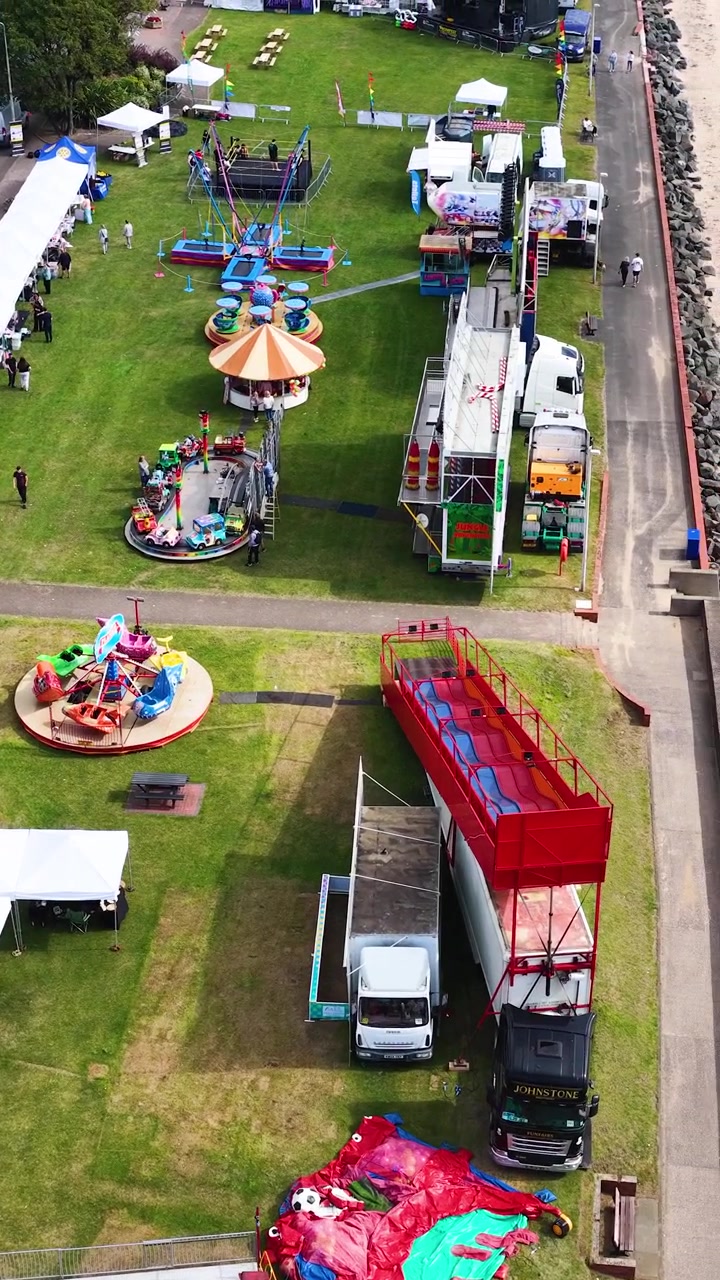 Drone svæver over tivoliforlystelser, køretøjer og havnemur på en solrig dag i Fife, Skotland.
