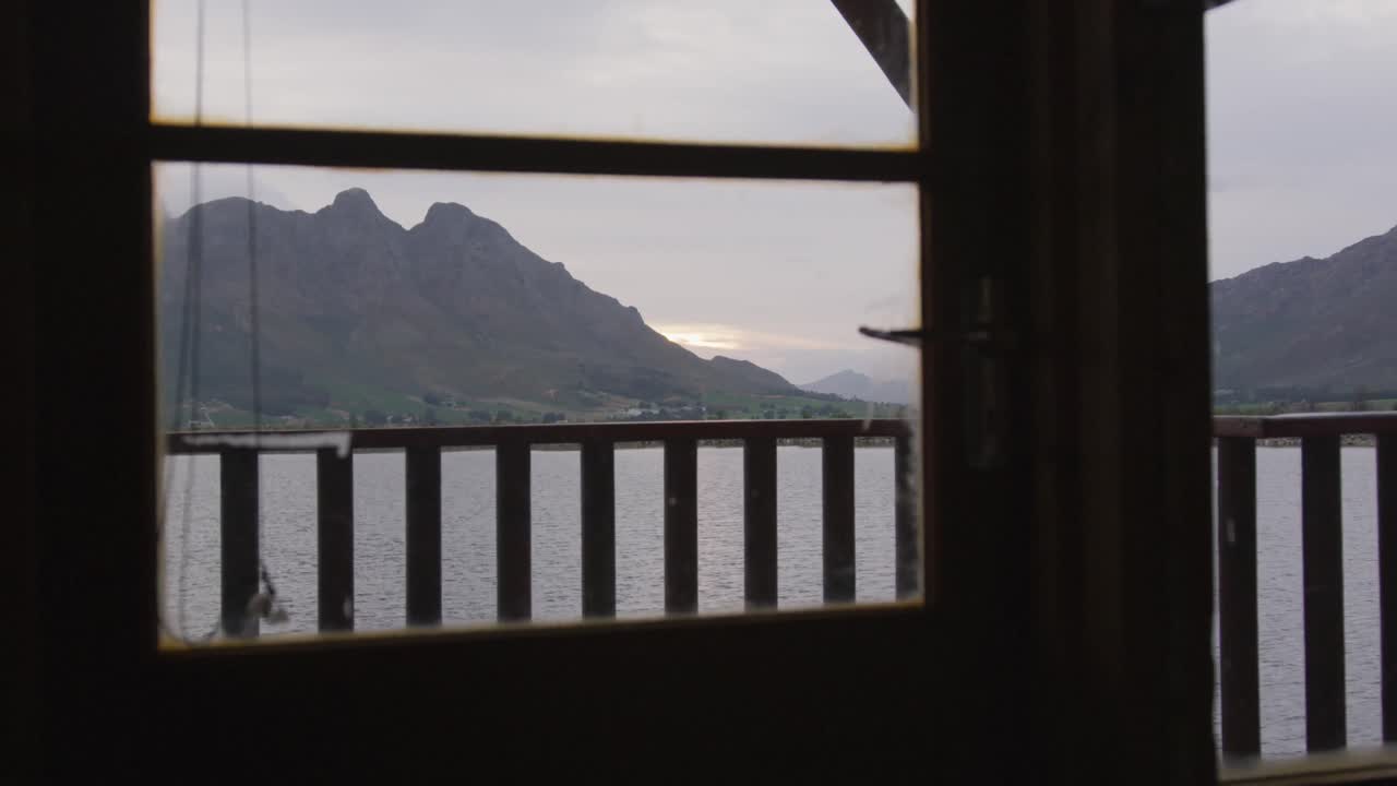 구름이 많은 날 해가 지는 동안 호수와 산의 전망이 로그 오두막 창문에서 보인다