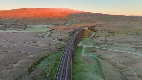 Geschwungene-Eingleisige-Eisenbahnbrücke-Mit-Beleuchtetem-Hügel-Im-Wintersonnenaufgang