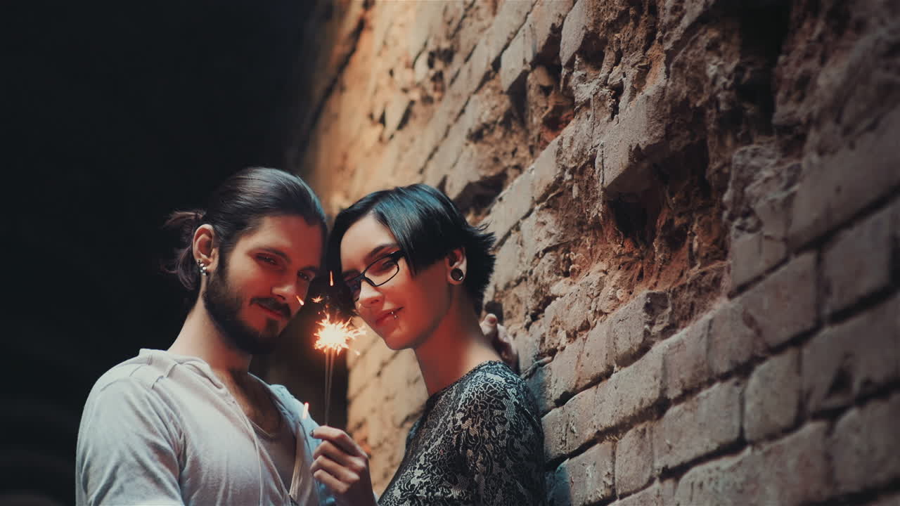 젊은 고딕 힙스터 부부는 벽돌 벽의 배경에 서 있는 스파클러를 존경합니다.