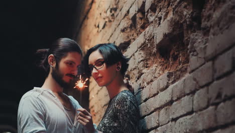 Young-Gothic-Hipster-Couple-Admires-The-Sparkler-Standing-Against-A-Background-Of-A-Brick-Wall-In-An