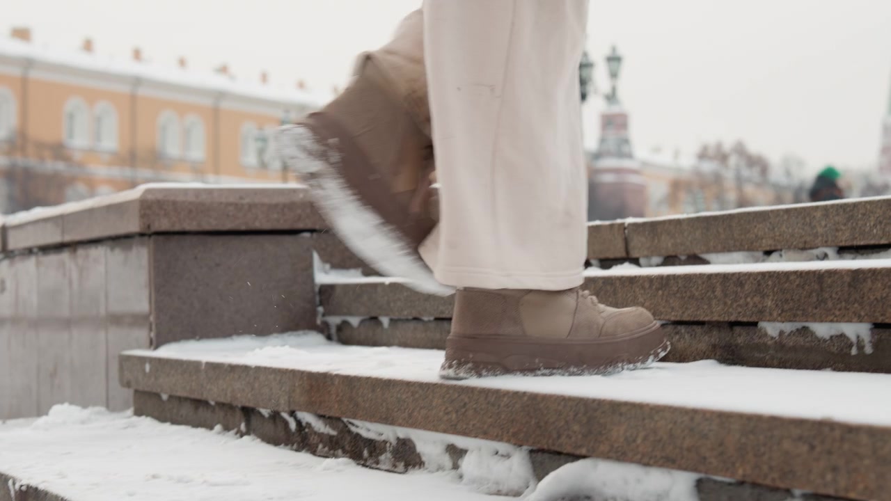 Low-angle close-up of lower legs and winter boots climbing outdoor stone steps dusted with fresh snow, showing lifted foot mid-step, scattered snow on treads, and subtle motion blur through cold