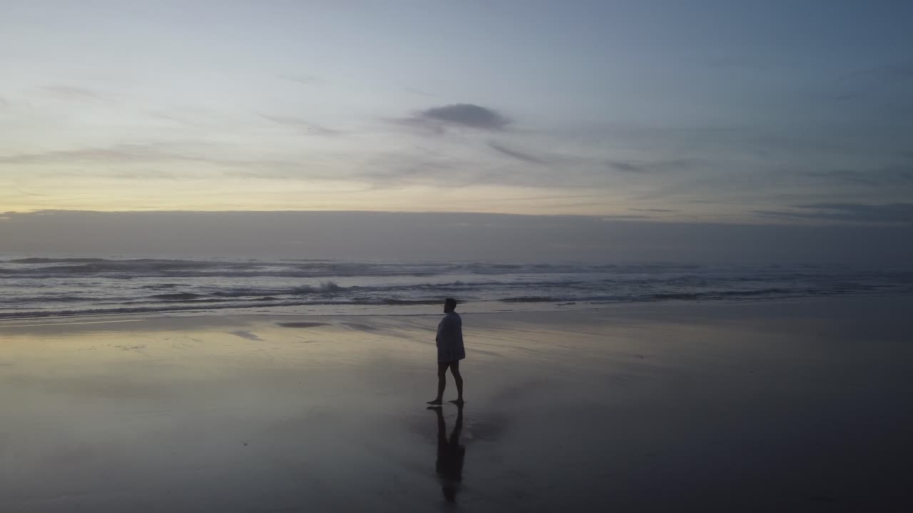 A tracking drone shot of a man walking towards the sea covered with a towel, ending with a shot of the ocean with a beautiful sunset as a background.