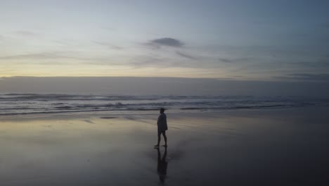 a tracking drone shot of a man walking towards the sea covered with a towel, ending with a shot of the ocean with a beautiful sunset as a background.
