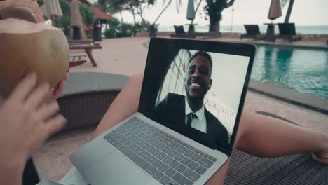 woman wearing white shirt and drinking from a coconut on video call with an coworker while sitting in the hammock in front of the pool in resort