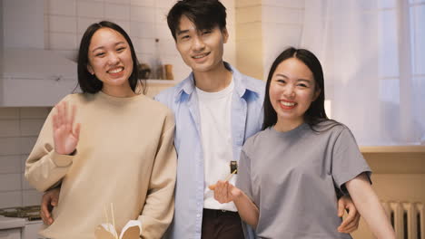 three japanese friends eating japanse food around the kitchen counter, then they look at camera, smile and wave