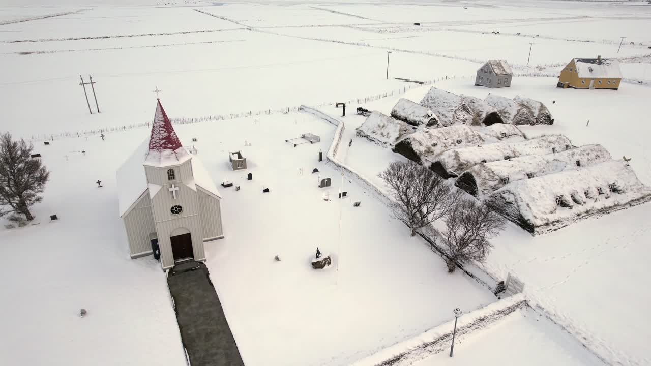 Aerial view of traditional turf houses and small church surrounded by thick snow in quiet countryside shows Icelandic heritage in peaceful frozen setting under soft winter light with empty fields