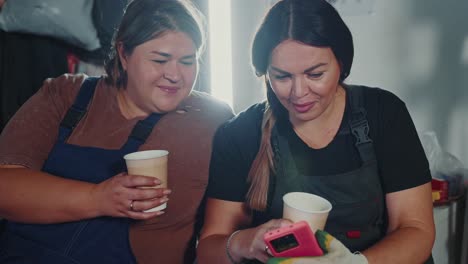 two women workers having a coffee break and discussing on smartphone