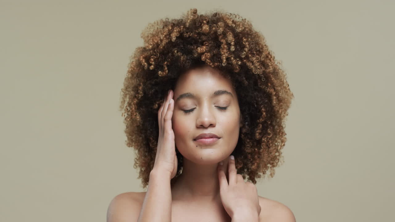Biracial woman with dark hair touching face on beige background with copy space, slow motion