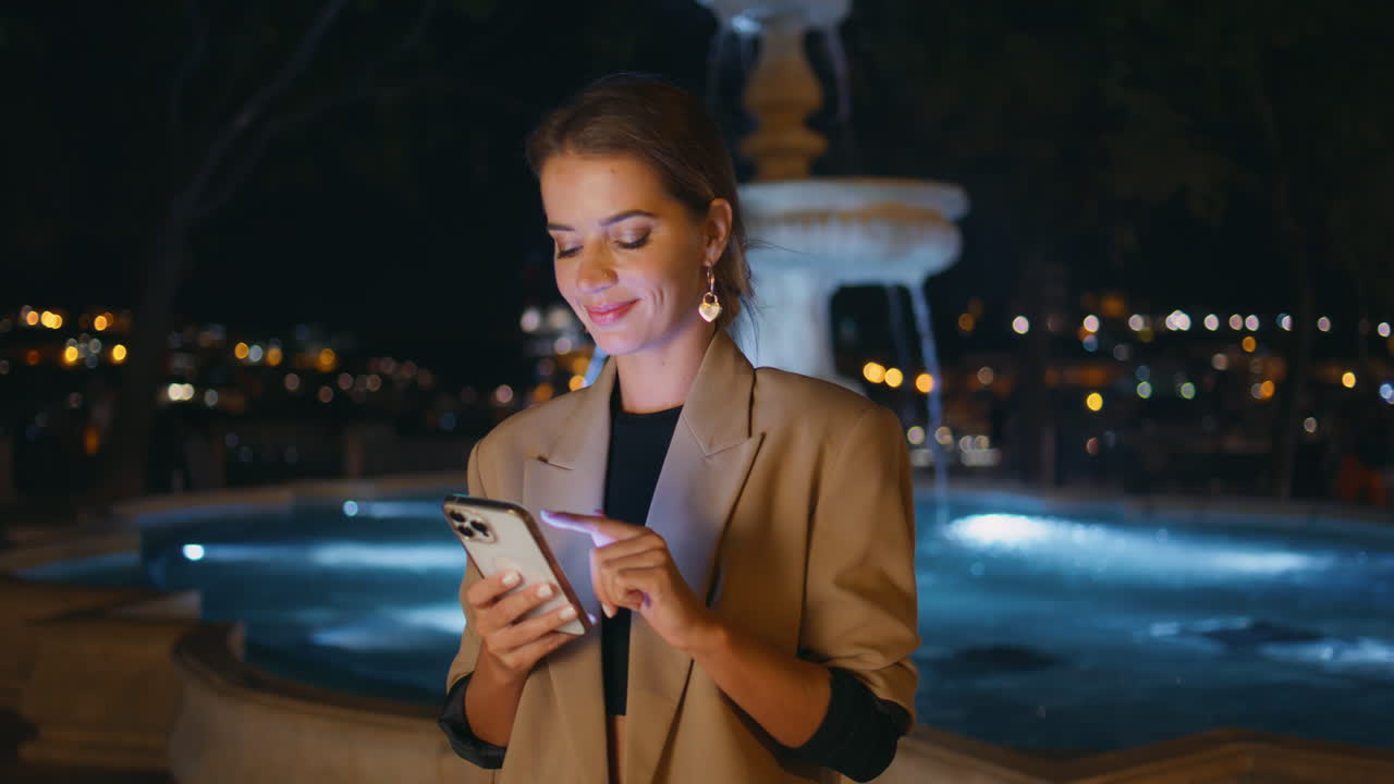 mujer sonriente desplazando el teléfono móvil en la ciudad de la noche. mujer elegante sosteniendo el celular