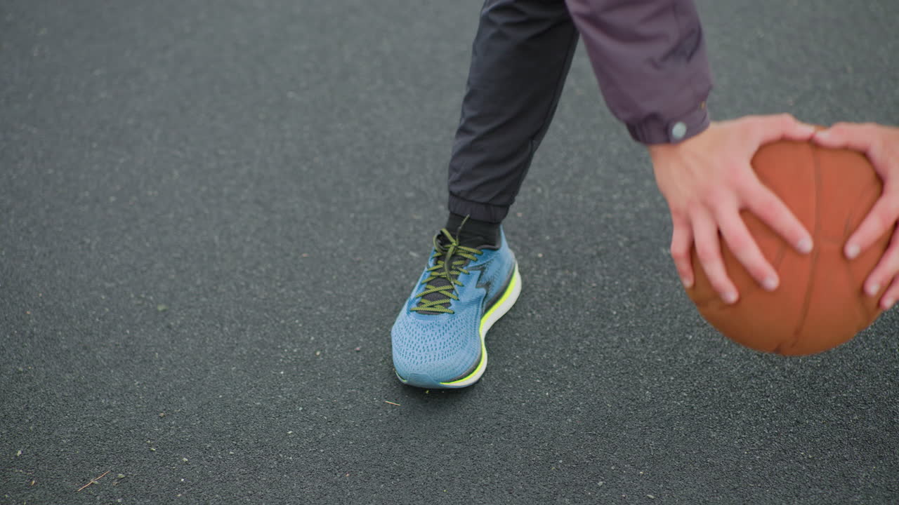 Primo piano di un palleggio di pallacanestro su asfalto, piede con scarpa da ginnastica che fa perno e mani che controllano la palla, superficie del campo all'aperto nuvoloso, pavimentazione ruvida, scena di allenamento mirata, manica della giacca casual visibile, slow