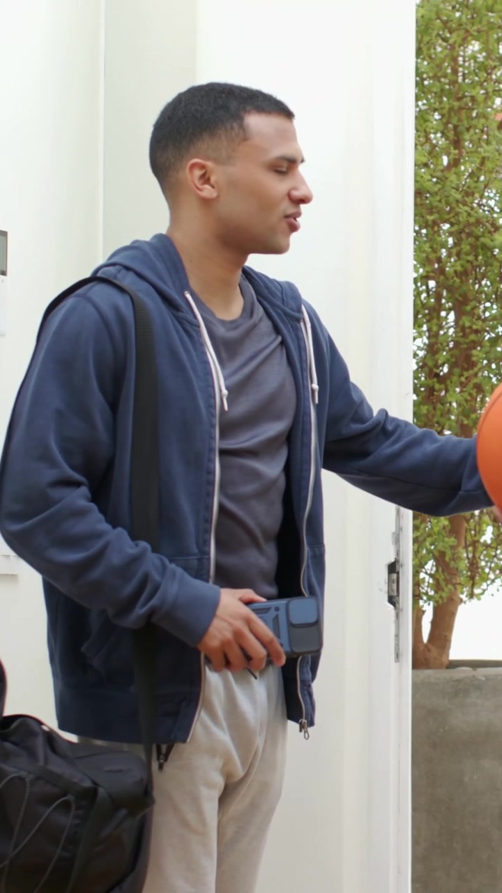 Vertical video: Stepping man in navy hoodie entering doorway receiving orange ball to join teammate