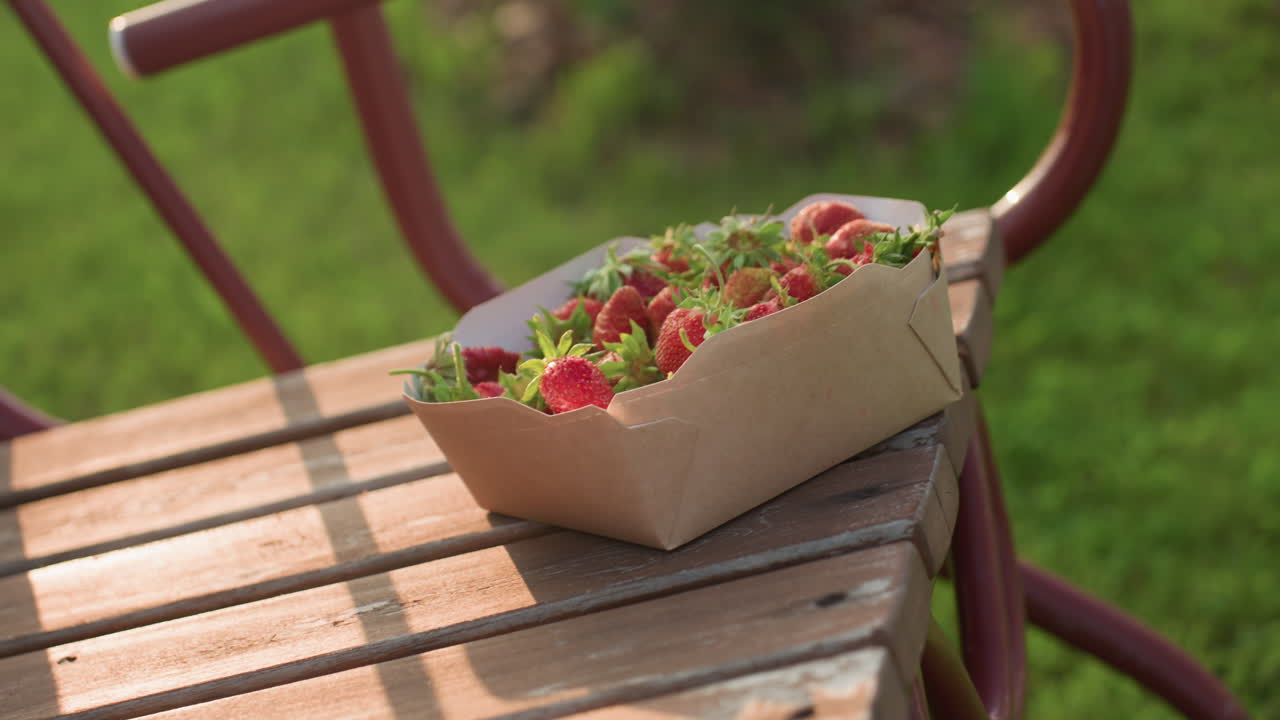 close up of fresh strawberry in cardboard container perched on wooden swing bench gently swaying with warm golden glow and blurred greenery background, sun rays creating soft pastoral scene