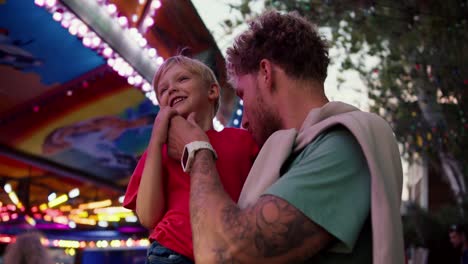 un niño rubio feliz con una camiseta roja toca la paja de su padre mientras está sentado en sus brazos en un parque de atracciones. padre e hijo juegan mientras el hijo se sienta en los brazos de papá en el parque de diversiones