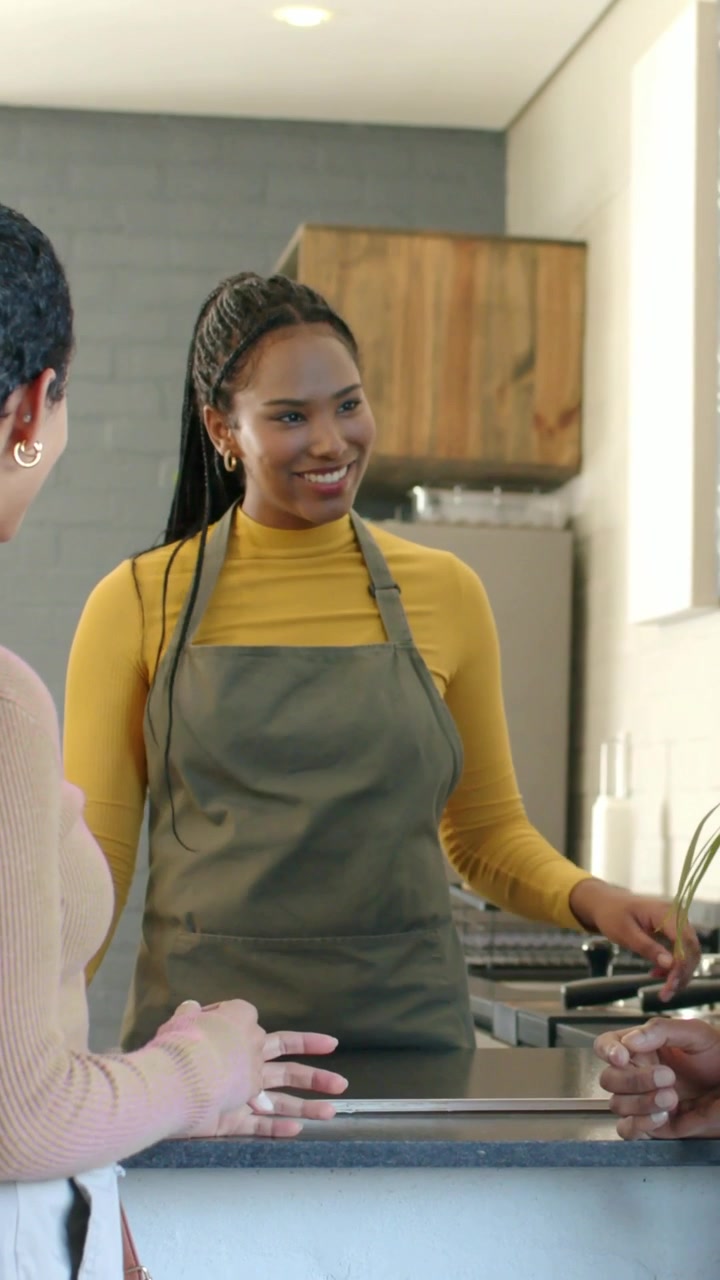 Staff member in mustard top, gray apron arriving behind counter smiling, leaning, serving customer