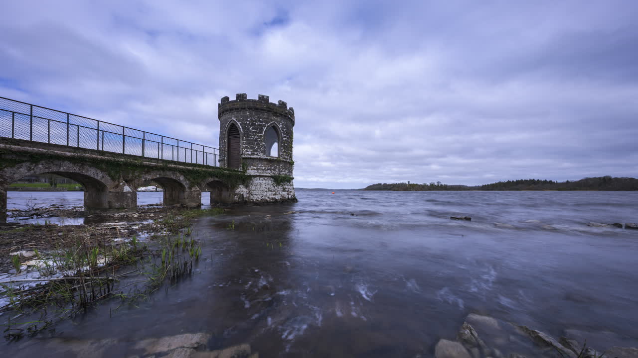 Zeitrafferaufnahme eines mittelalterlichen Turms an einem See mit Uferfelsen im Vordergrund und einer mittelalterlichen Burg in der Ferne an einem bewölkten Tag im County Roscommon in Irland