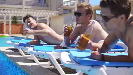 group of young friends drinking beer and relaxing lying on the coaches by the swimming pool. slow motion shot