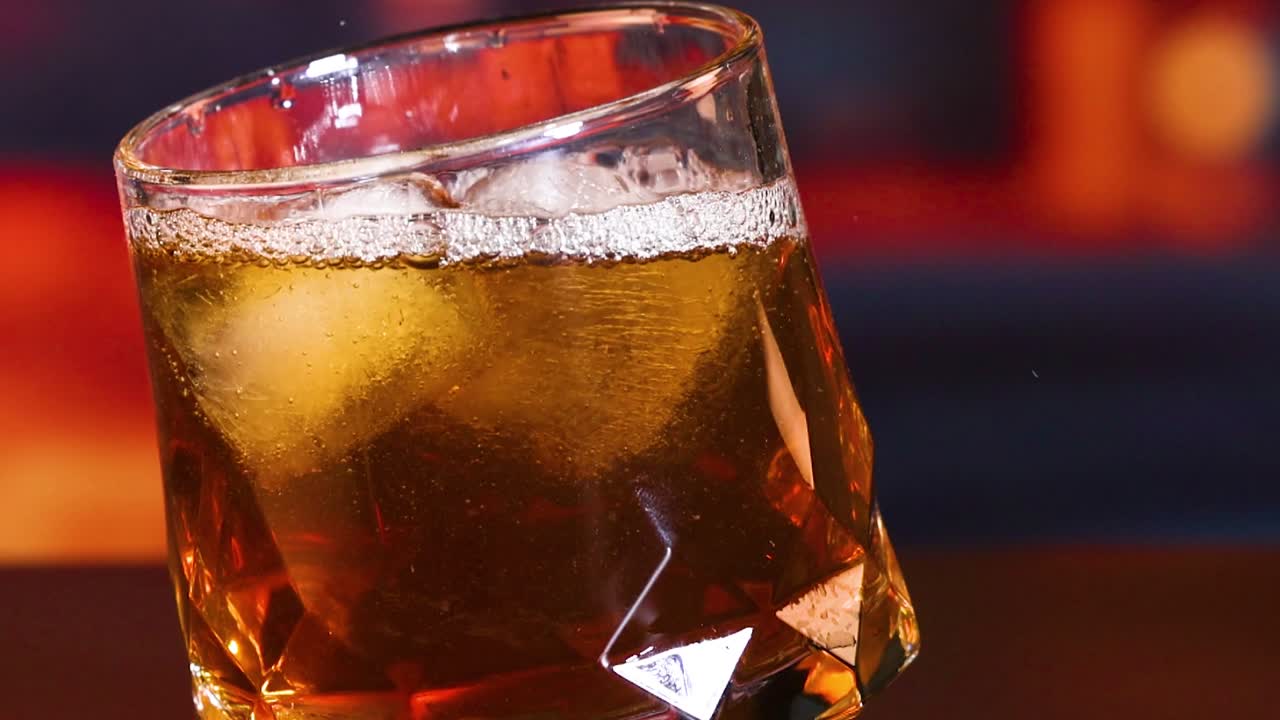 Close-up of a golden liquid being poured into a glass with ice cubes, creating bubbles and reflections.