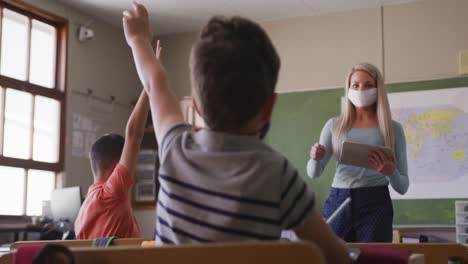 female teacher wearing face mask pointing towards a boy raising his hands in class at school