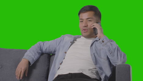 studio shot of young man sitting on sofa talking on mobile phone against green screen 1