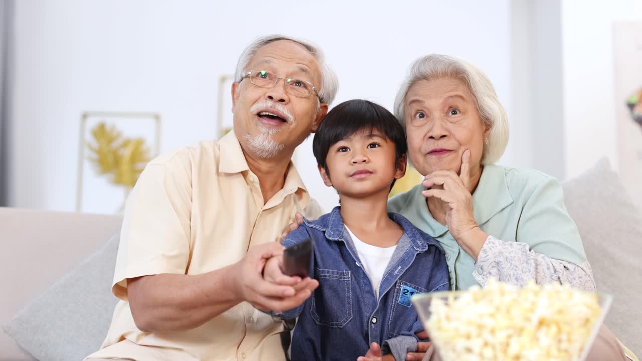 Grandparents and grandson laughing together, sharing popcorn while watching a movie in cozy living room