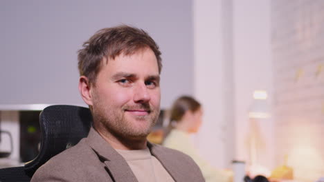 portrait of a smiling businessman sitting on office chair and looking at camera while two employees working behind him 1