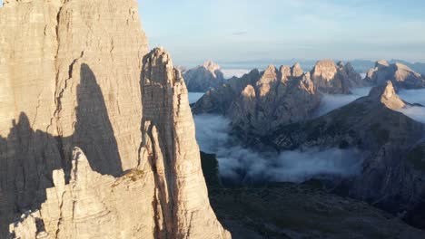 cinematic drone shot showing tre cime spires in dolomite mountains during a golden sunrise - clouds covering valley in background - panoramic shot