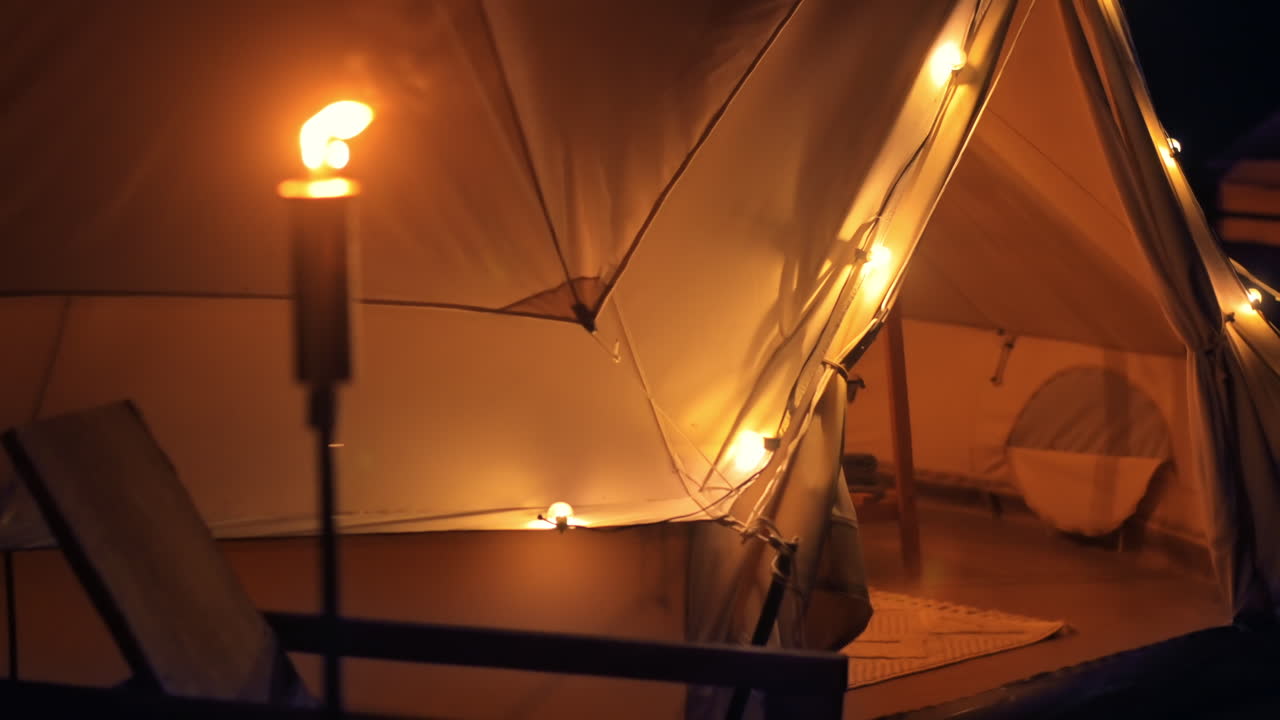 Tenda con torcia accesa, lampade e sedia di legno in un sito di glamping, notte