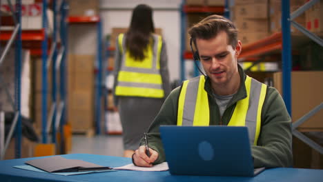 trabajador masculino con auriculares sentado en un escritorio trabajando en una computadora portátil en un almacén ocupado