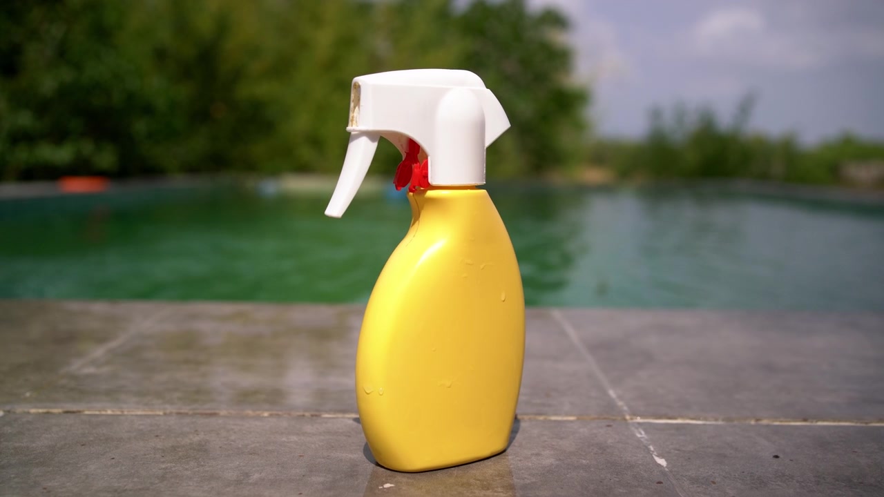 Una bottiglia spray gialla di crema solare si trova sul bordo della piscina mentre l'acqua increspa dietro di essa, Ripresa a mano.