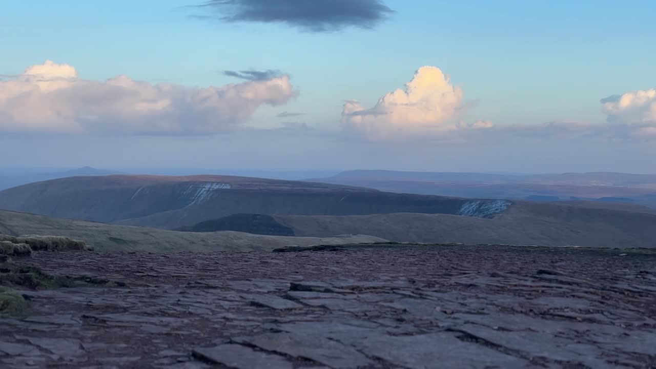 Altopiano sommitale roccioso a Pen y Fan con ampio terreno in pietra e cime ondulate distanti sotto nuvole soffici e una calda luce serale che crea una vista panoramica del paesaggio montano.