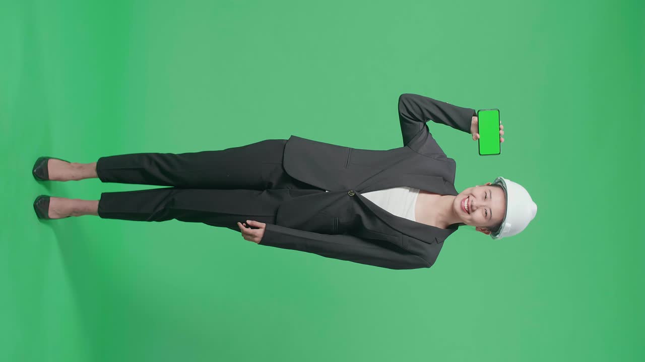 Full Body Of Smiling Asian Female Engineer With Safety Helmet Showing Green Screen Smartphone In The Green Screen Studio