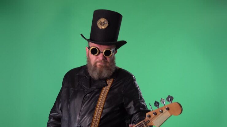 A bearded man in a long leather coat, an Irish style hat and original glasses plays the red guitar. Rock musician plays bass guitar in studio on green screen chroma key. Slow motion. Close up