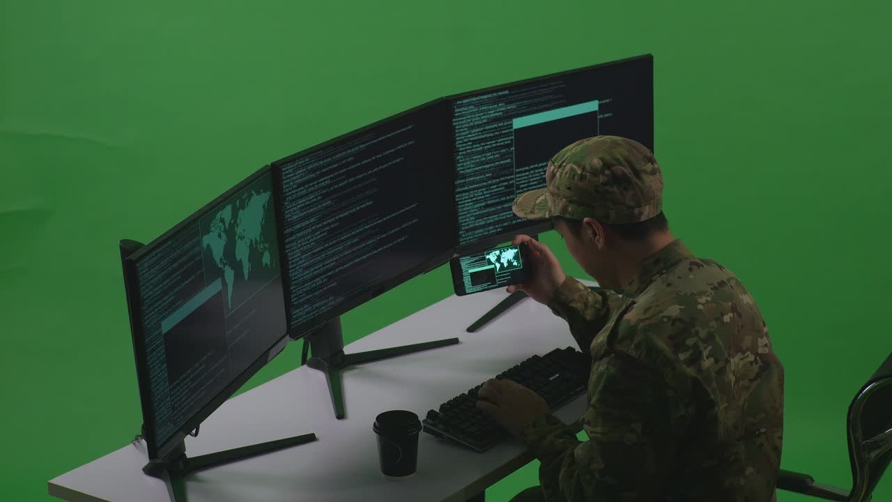 Side View Of Asian Man Soldier Developer Working With Mobile Phone While Write Code With Multiple Computer Screens In Green Screen Studio