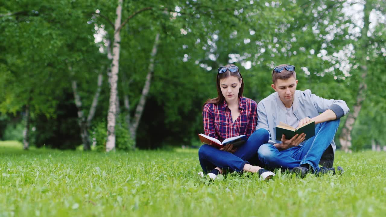 Joven y una mujer leyendo un libro en el parque, se sientan en un césped verde, vídeo HD