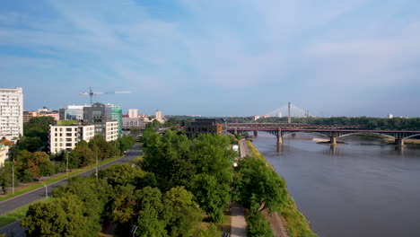 Aerial-flight-over-path-beside-Vistula-River-and-bridge-in-city-of-Warsaw,-Poland-during-sunny-day