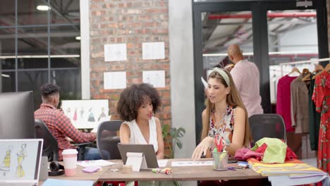 Two-happy-diverse-female-designers-sitting-and-discussing-designs-at-fashion-studio,-in-slow-motion