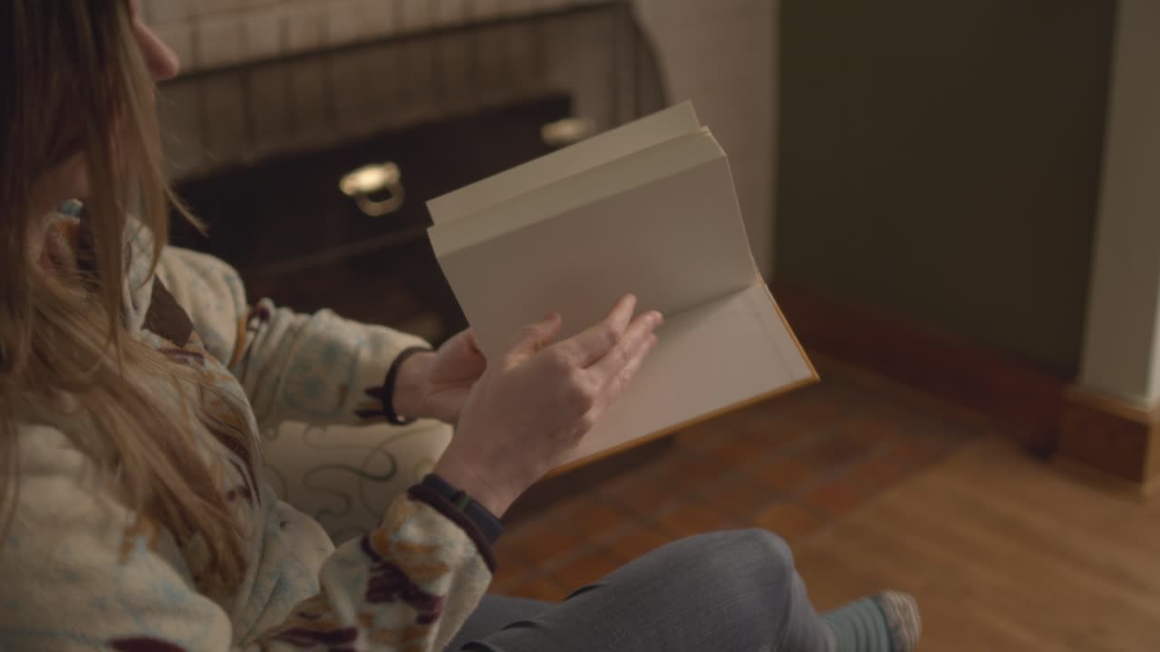 Over the shoulder close up of a woman flipping through the pages of book. A cozy living room in the house. She flips through the entire contents of the book. Shot in 60 frames for slow motion