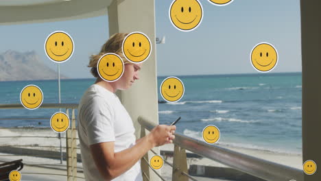 animation of social media emojis over man using smartphone by seaside