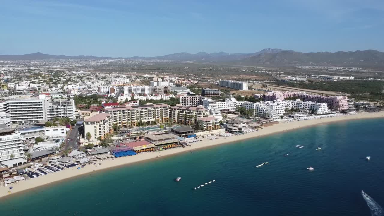 toma aérea sobre la maravillosa costa de la playa de medano en cabo san lucas con vistas al mar azul con barcos flotantes, edificios de hoteles y un hermoso paseo marítimo durante un viaje a méxico