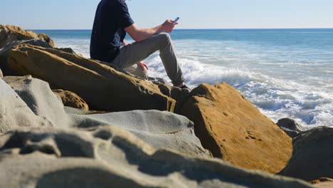 a man sitting down texting on his smartphone as california ocean waves crash and spray against the rocky shore in slow motion