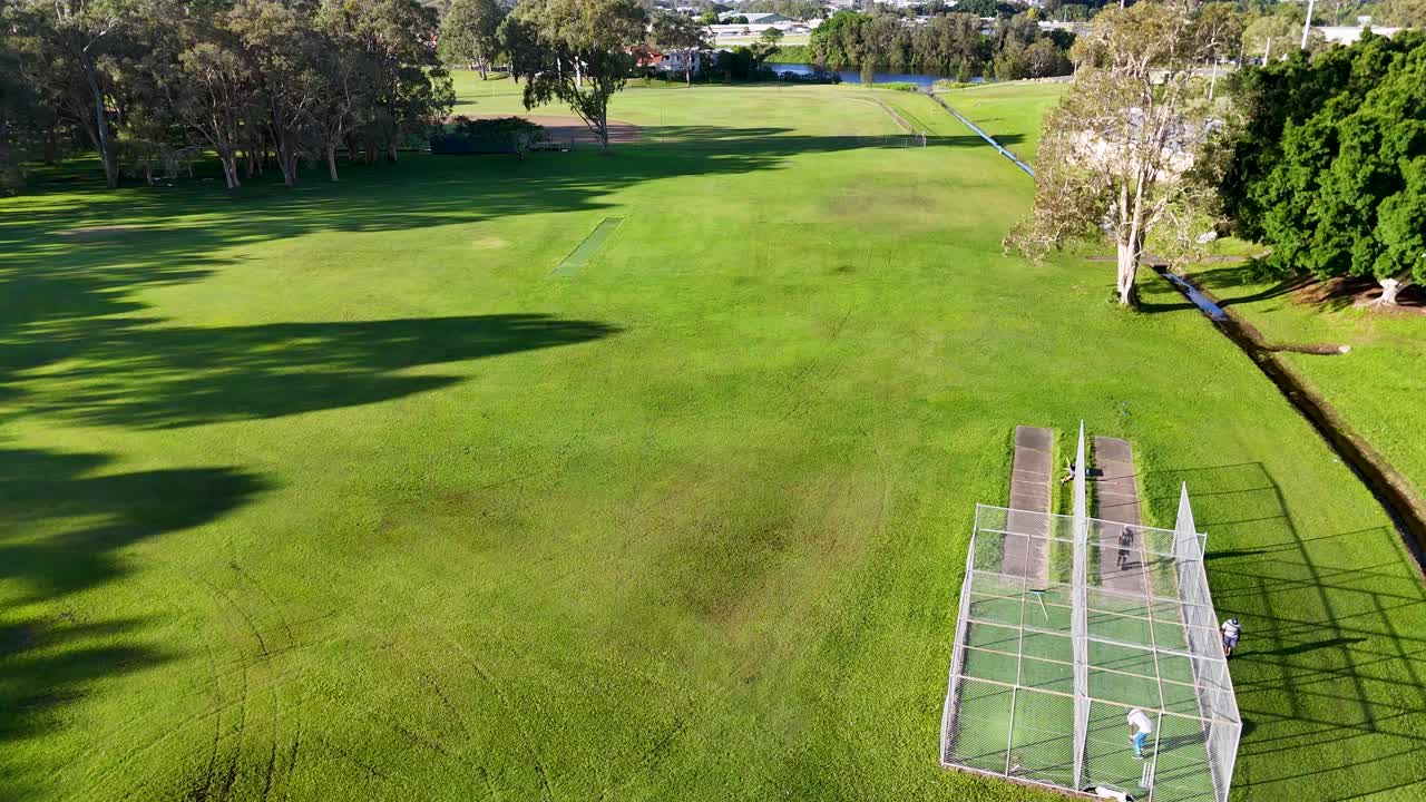 Aerial footage of cricket practice nets in a lush park setting, captured in bright daylight with clear skies