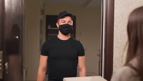 pizza provider in black uniform and protective mask and gloves hands over paper boxes to client in apartment house