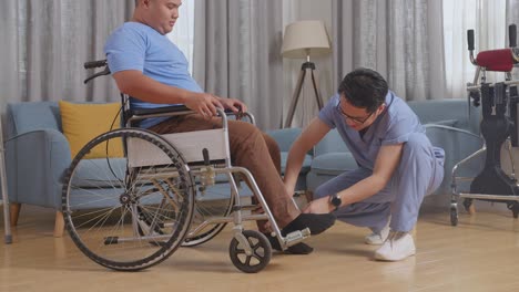 Side View Of Asian Male Nurse Helping A Patient In Wheelchair Putting Feet On Footrest And Pushing Him To Another Room After A Physical Therapy At Home