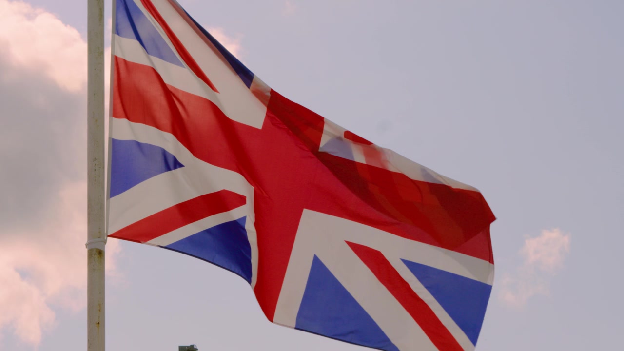 Cờ Liên hiệp Anh (Union Jack) bay phấp phới trong gió với chuyển động chậm, nền trời xanh nắng và mây. Cảnh quay khái niệm yêu nước, dân tộc 4K