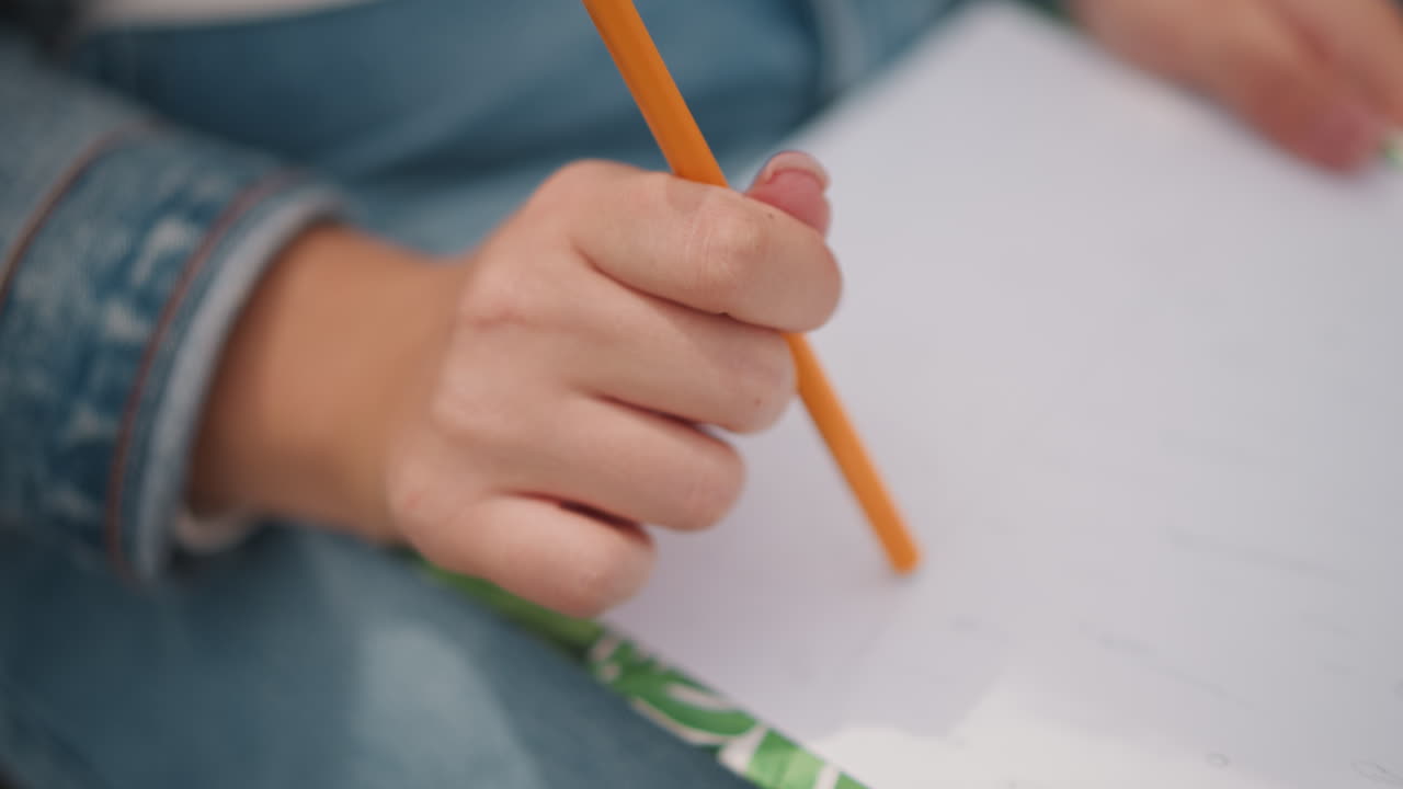 Child hand holding pencil on paper intimate close up of young student wearing denim jacket gripping orange pencil above patterned notebook careful handwriting practice during quiet homework session